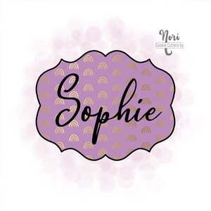 Peut inclure: Emporte-pièce violet et or avec un motif arc-en-ciel décoratif et le nom "Sophie" en écriture cursive noire. Nori Cookie Cutters by.