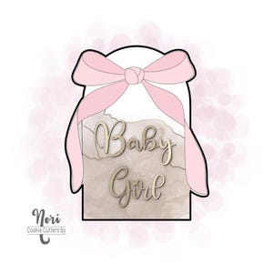 May include: A pink and white cookie cutter in the shape of a gift box with a bow on top. The box has a marbleized design and the words "Baby Girl" written in gold.