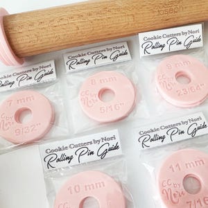 May include: Set of six pink plastic rolling pin guides for cookie cutters. Each guide has a different size marked in millimetres and inches. The guides are packaged individually in clear plastic bags with a label that reads "Cookie Cutters by Nori Rolling Pin Guide".