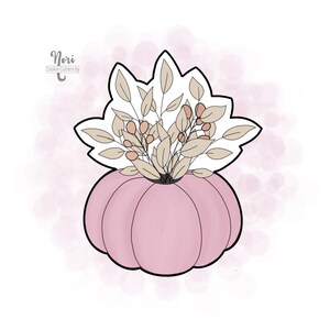 May include: A pink pumpkin cookie cutter with a floral arrangement on top. The floral arrangement includes beige leaves and small pink berries. The cookie cutter has a black outline and the text "Nori Cookie Cutters by" in the upper left corner.