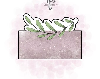 May include: A cookie cutter in the shape of a rectangular box with a sprig of green leaves on top. The cookie cutter is on a pink and white background with a white text overlay that says "Nori Cookie Cutters by C".