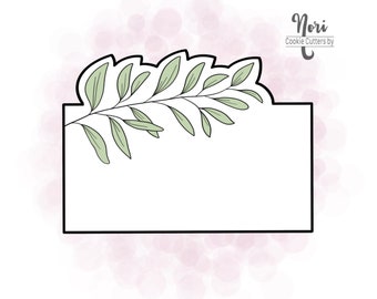 May include: A black and white cookie cutter in the shape of a rectangle with a leafy branch design at the top. The text "Nori Cookie Cutters by" is in the top right corner of the image.