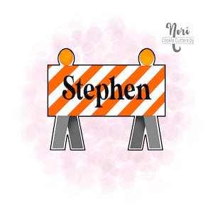 May include: A cookie cutter in the shape of a road construction barrier. The barrier is white with orange diagonal stripes and the name "Stephen" in black. Two orange lights sit atop the barrier, with gray legs supporting it.