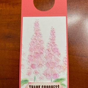 May include: Pink floral design on a white background with the text "THANK GOODNESS we haven't hit that age where we know better yet". The design is on a pink card with a hole at the top for hanging.