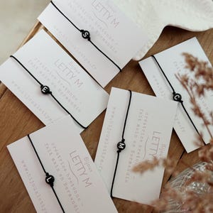 Peut inclure: Plusieurs bracelets noirs avec des breloques lettres, présentés sur des cartes blanches avec le texte "LETTY M Bijoux" et les coordonnées du site web. L'ensemble est disposé sur une surface en bois.