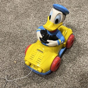 May include: A vintage Donald Duck pull-along toy car. The yellow car has red wheels and a blue base. Donald Duck is wearing a blue sailor suit and hat. The toy has a white cord attached.