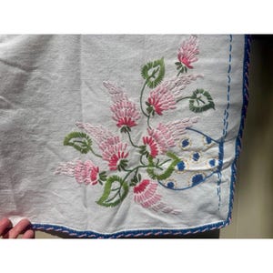 May include: White linen textile with embroidered pink and green floral design. The embroidery features detailed pink flowers with green leaves and stems. The edge is trimmed with a blue and pink stitched border.