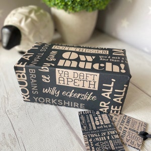 May include: A black gift box with brown text and a white sheep figurine in the background. The text on the box reads "Doit Thissen", "Ya Daft Apeth", "Willy eckerslike", "Yorkshire", and "Much!"
