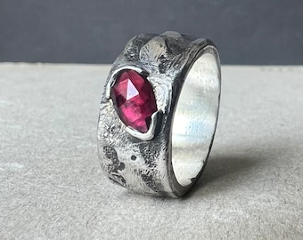 May include: A silver ring with a textured band and a deep pink gemstone set in the center.