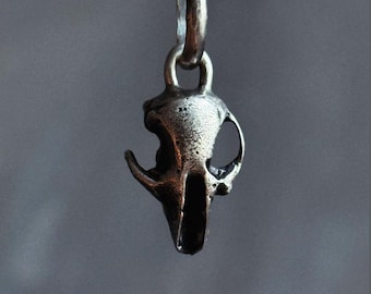 May include: A silver-colored pendant in the shape of a small animal skull, suspended from a silver chain. The skull has detailed features, including eye sockets and a nasal cavity. The background is a blurred gray.