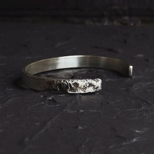 Könnte beinhalten: Ein silbernes Armreif-Armband mit einem strukturierten, robusten Design an einem Ende. Das offene Armband wird vor einer dunklen, strukturierten Oberfläche präsentiert, die seinen metallischen Glanz und seine einzigartige Ästhetik hervorhebt. Ein Schmuckstück.