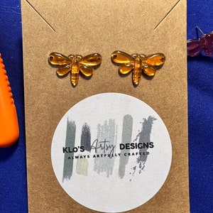 Op de afbeelding: Een paar amber glazen libellen oorbellen op een bruine kaart met de tekst "KLO'S Artsy DESIGNS ALWAYS ARTFULLY CRAFTED".
