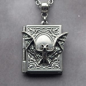 May include: Silver pendant necklace. The pendant is a rectangular locket with a bat design. The bat has outstretched wings. The necklace has a chain with small, round links. The locket is approximately 2.5 cm in height.