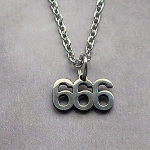 May include: A silver necklace with a chain and a pendant. The pendant displays the number "666" in a stylized font. The necklace is set against a grey background.