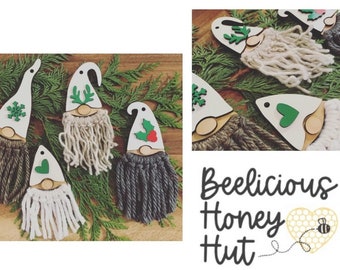 May include: Five wooden gnome ornaments with different colored yarn beards and green felt accents. The gnomes are decorated with a snowflake, antlers, a heart, and holly. The ornaments are displayed on a bed of green pine branches. The text "Beelicious Honey Hut" is visible in the bottom right corner of the image.