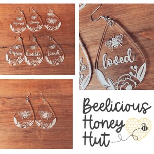 May include: A set of six clear acrylic teardrop earrings with white floral designs and bee illustrations. Each earring has a different word inscribed on it: "inspired", "strong", "happy", "humble", "loved", and "loved". The earrings are displayed on a wooden surface. The image also includes a logo for "Beelicious Honey Hut" with a bee and honeycomb design.