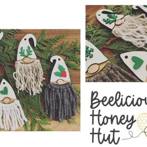 May include: Five wooden gnome ornaments with different colored yarn beards and green felt accents. The gnomes are decorated with a snowflake, antlers, a heart, and holly. The ornaments are displayed on a bed of green pine branches. The text "Beelicious Honey Hut" is visible in the bottom right corner of the image.