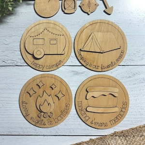 May include: Set of six wooden round ornaments with camping-themed designs. The ornaments feature a camper, tent, campfire, s'mores, compass, and lantern. The text on the ornaments reads "happy camper", "camping is our favorite season", "starry nights & bonfires", "making s'more memories", and "camping".
