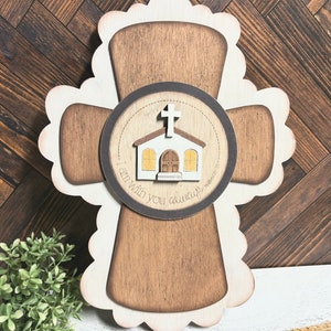 May include: A wooden cross with a scalloped edge, painted in a light brown color. The center of the cross features a round, brown plaque with a white church and a cross on top. The plaque has the text "I am with you always" written around the church.