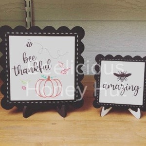 May include: Two small, black framed signs with scalloped edges. One sign says "bee thankful" with a pumpkin and bee graphic. The other says "amazing" with a bee graphic. Both signs are on wooden easels.