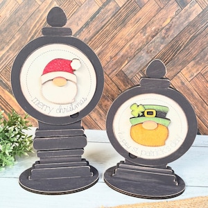 Op de afbeelding: Twee houten kerst- en St. Patricks Day-decoraties. Elke decoratie heeft een rond, zwart omlijst bord met een feestelijk personage en een boodschap. De kerstdecoratie heeft een kerstman met de boodschap "merry christmas". De St. Patricks Day-decoratie heeft een kabouter met de boodschap "happy st. patrick's day".