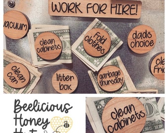 Pode incluir: Uma placa de madeira com o texto "WORK FOR HIRE!" e vários tokens redondos de madeira com diferentes tarefas escritas neles, como "limpar os armários", "dobrar as roupas", "escolha do pai", "limpar a lata", "caixa de areia", "lixo de quinta-feira", e "limpar os armários".