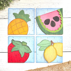 Peut inclure: Quatre carrés en bois avec un fond bleu et des étoiles blanches. Chaque carré présente un fruit différent : un ananas, une tranche de pastèque, une fraise et deux citrons.