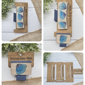 May include: Wooden display stand with three slots for honey jars. Each slot has a cutout for a jar and the text "Beelicious Honey Farms" printed on the wood. The stand is made of natural wood with a light brown colour.