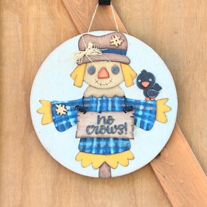 Puede incluir: Un letrero redondo de madera con un espantapájaros pintado que lleva una camisa a cuadros azul y blanca y un sombrero marrón. El espantapájaros sostiene un letrero que dice "no crows!" y tiene un pequeño pájaro negro posado en su brazo.