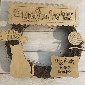 May include: Wooden cat-themed wreath with a welcome sign that says "Welcome to our home", a cat silhouette, a fishbone, a ball of yarn, and a sign that says "My kids have paws".