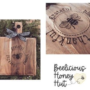 Puede incluir: Tabla de cortar de madera con una cinta a cuadros en blanco y negro. La tabla está grabada con una abeja y las palabras "thankful", "grateful" y "blessed".