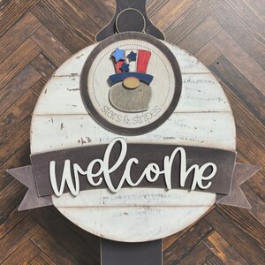 May include: A white wooden round sign with a brown banner that says "welcome". The sign has a brown circle with a red, white, and blue gnome wearing a hat with stars and stripes. The circle says "stars & stripes".