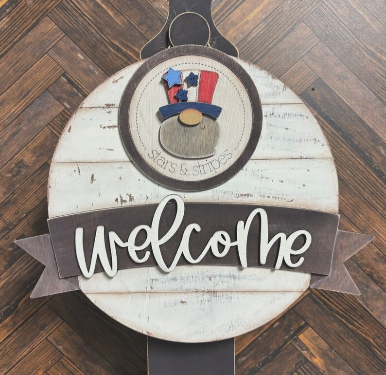 May include: A white wooden round sign with a brown banner that says "welcome". The sign has a brown circle with a red, white, and blue gnome wearing a hat with stars and stripes. The circle says "stars & stripes".