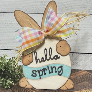 Puede incluir: Decoración de primavera de madera con forma de conejo con un lazo de cuadros pastel, paja y las palabras "hello spring" en la parte delantera.
