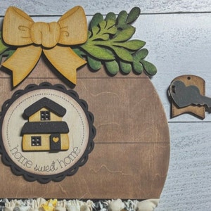 May include: Wooden home sweet home sign with a yellow bow, green leaves, and a small house with a heart on the door. The sign is on a white wood background.