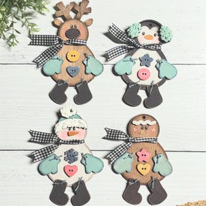 Może przedstawiać: Cztery drewniane ozdoby świąteczne: renifer, pingwin, bałwan i piernik. Każda ma czarno-białą kratowaną wstążkę, rękawice i guzik. Renifer ma rogi, pingwin ma nauszniki, bałwan ma kapelusz, a piernik ma uśmiech.
