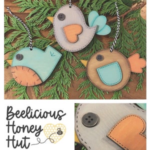 May include: Three wooden bird ornaments with painted details. The birds are blue, brown, and grey with orange accents. The ornaments are hanging from twine and have a stitched look. The text "Beelicious Honey Hut" is visible in the image.