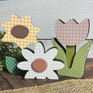 May include: Three wooden flower decorations with gingham patterns. A yellow sunflower, a pink tulip, and a white daisy.