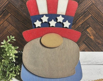 May include: A wooden gnome wearing a red, white and blue hat. The hat has white stars on a blue background. The gnome's beard is brown and the hat is shaped like a traditional American flag.