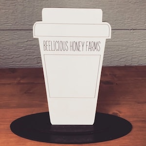 May include: White wooden cutout of a coffee cup with the text "Beelicious Honey Farms" on the side. The cup is standing on a black circular base.