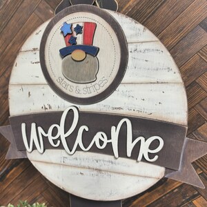 May include: A round wooden welcome sign with a whitewashed finish. The sign features a red, white, and blue gnome wearing a top hat with stars and stripes. The sign also has the words "stars & stripes" and "welcome" in black lettering.