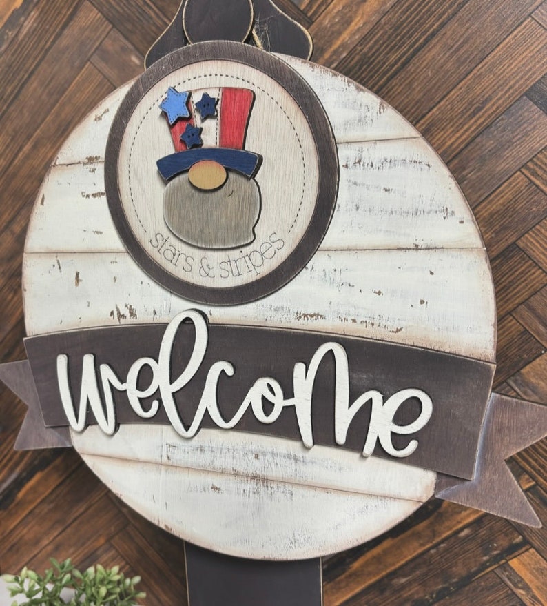 May include: A round wooden welcome sign with a whitewashed finish. The sign features a red, white, and blue gnome wearing a top hat with stars and stripes. The sign also has the words "stars & stripes" and "welcome" in black lettering.
