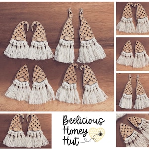 May include: A set of 10 wooden gnome earrings with white fringe. The gnomes are painted with a light brown color and have a white beard. The earrings are handmade and have a rustic look. The earrings are labeled "Beelicious Honey Hut".