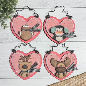 May include: Four heart-shaped wooden ornaments with scalloped edges. Each ornament features a different winter character: a gingerbread man, a penguin with earmuffs, a reindeer, and a moose. Each character has a black and white checkered scarf and a black wire hanger.