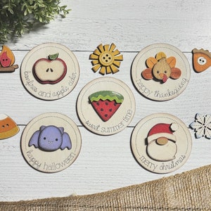 Könnte beinhalten: Neun runde Holzornamente mit verschiedenen Feiertagsmotiven. Jedes Ornament zeigt eine kleine, bunte Illustration und Text. Die Ornamente beinhalten ein Lagerfeuer, einen Apfel, eine Wassermelone, einen Truthahn, eine Schneeflocke, eine Fledermaus, den Weihnachtsmann, einen Kuchen und eine Sonnenblume.