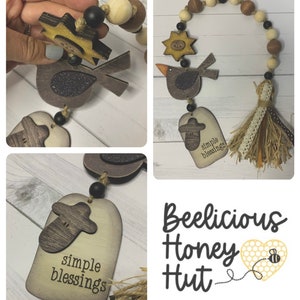 May include: A wooden door hanger with a brown bird, a star, and a beehive. The hanger has a brown and white tassel and the text "simple blessings" on a wooden tag. The hanger is decorated with wooden beads and a burlap ribbon.