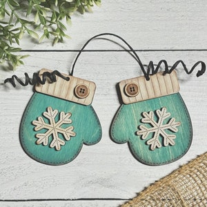 May include: Two teal blue wooden ornaments shaped like mittens with a white snowflake design and brown button accents. The ornaments are connected by a black string.