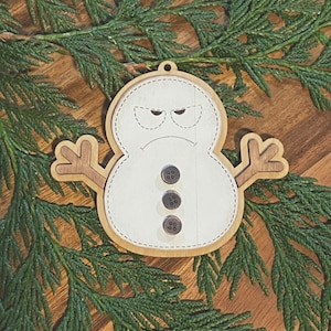 May include: A wooden snowman ornament with a grumpy expression and three black buttons. The ornament is painted white and has a brown wooden base.