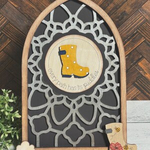 Peut inclure: Une pancarte en bois avec un fond noir et un motif de mandala argenté. La pancarte a une découpe circulaire avec une botte de pluie jaune à pois blancs et le texte "every path has its puddles".