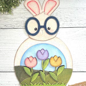 May include: A white wooden bunny with blue glasses holding a circle with three colorful tulips inside. The tulips are pink, purple, and yellow. The bunny is standing on a green grassy field.